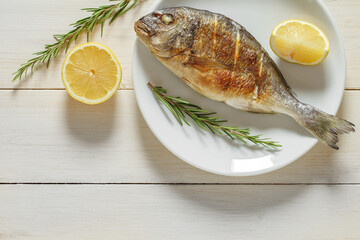 Dorado, cooked, grilled fish fried, rosemary, cut lemons, source of omega 3, on plate, on white wooden board background, top view, space to copy text