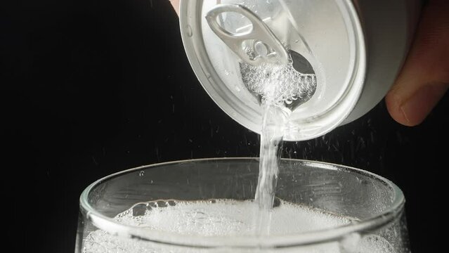 The scene slowly rotates, and soda is slowly poured out of a can on a black background, close-up.