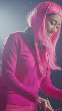 Vertical video: Performer mixing electronic sound with techno, enjoying party with fans, standing at dj table performing at party in club at night. Asian musician with pink hair performing music using