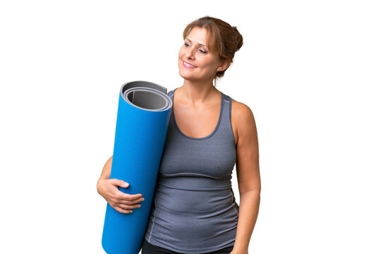 Middle-aged sport woman going to yoga classes while holding a mat over isolated background looking to the side and smiling