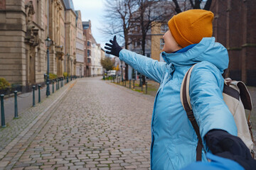 winter travel to Dusseldorf, Germany. young Asian tourist in blue jacket and yellow hat (symbol of Ukraine) making follow me pose on old town or Altstadt. Popular center of Rheinland and Westphalia