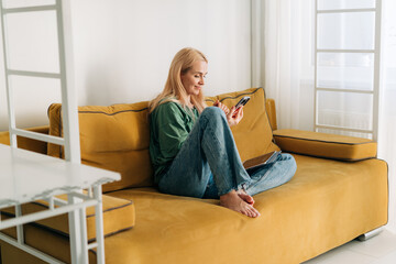 Caucasian middle aged woman scrolling internet in mobile phone while sitting on yellow sofa at home.