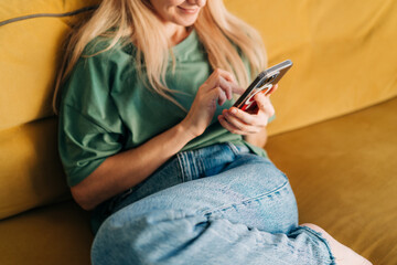 Unrecognizable woman browsing media on mobile phone while sitting on yellow sofa at home.
