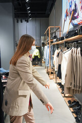  Beautiful woman is wearing protective face mask in shopping center. Girl trying on clothes. Concept of virus protection. Fashion, beauty and shopping concept.