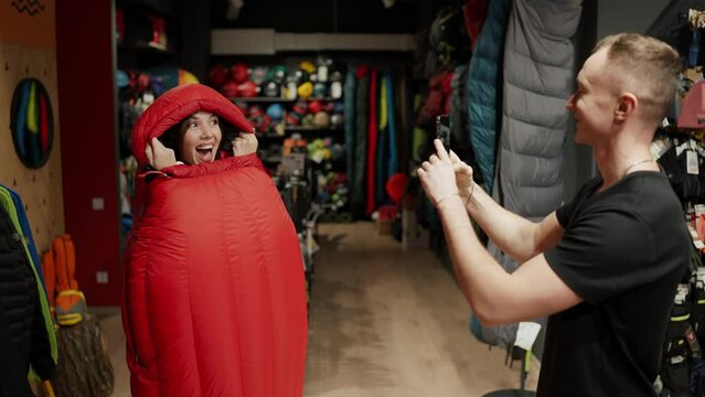 A smiling couple purchase in sportswear store taking pictures inside the sleeping bag