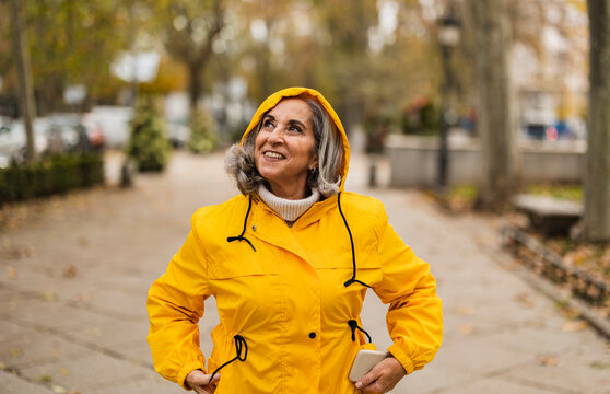 Thoughtful senior woman wearing yellow raincoat standing at footpath