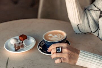Fototapeta na wymiar A cup of fresh cappuccino coffee in the hands of a woman on a fashionable background of a white marble table, next to a plate with sweets. Coffee addiction