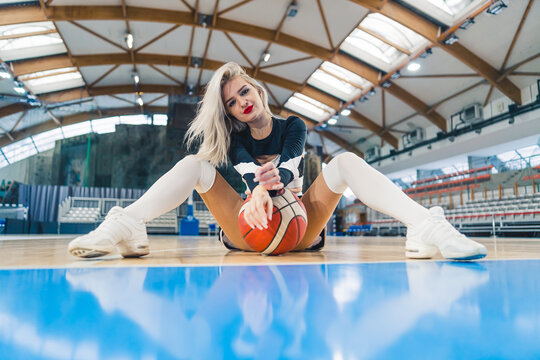 Low angle shot of a blond cheerleader sitting on the floor with a basketball between her legs. Sport concept. High quality photo