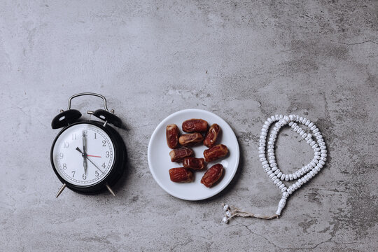 Date fruits on plate, alarm clock showing iftar time and prayer beads on gray background 