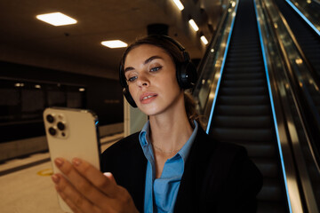 Young woman in headphones using mobile phone while standing by the escalator
