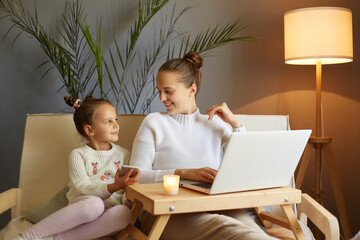 Horizontal shot of attractive woman in white sweater sitting on sofa with her daughter and using laptop computer, talking and enjoying time together at home at weekend.