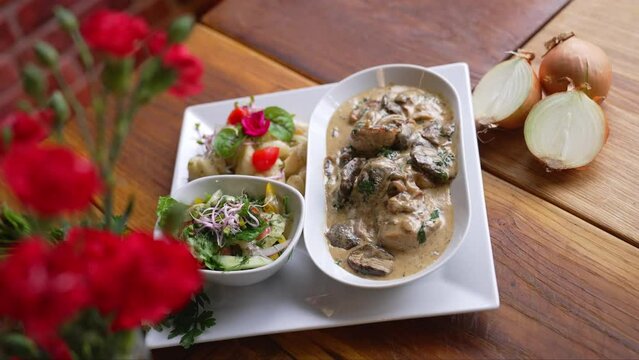 Traditional Polish dish - poledwiczki - tenderloin strips in mushroom sauce served with salad and potatoes. Blurred red flowers in the foreground. High quality 4k footage