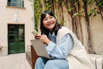 Smiling asian girl using mobile phone and laptop while sitting outdoors