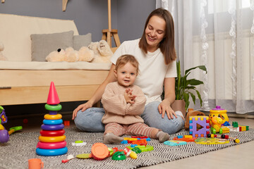 Indoor shot of happy female and her cute daughter sitting on floor and spending time together, kid clapping hands, family posing among lots of colorful toys.