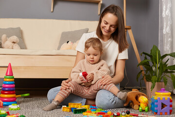 Image of smiling mother and child playing on the floor in the nursery with building a tower of colored blocks. Educational activities of children with parents at home.