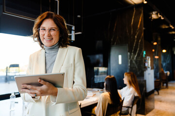 Mature woman in jacket smiling and using tablet during offline meeting