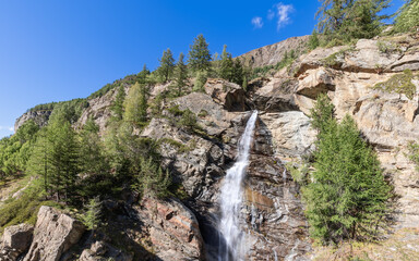 Fototapeta na wymiar Straight view of water stream of Lillaz waterfall (Cascate di Lillaz) on granite ledges of rock with green pine trees in Gran Paradiso National Park. Aosta valley, Italy