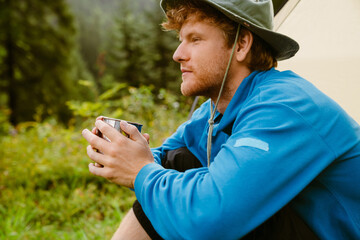 Fototapeta premium Young white tourist man drinking tea while sitting at tent in forest