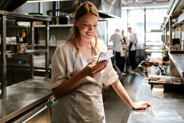 Young blonde woman using cellphone while working in restaurant kitchen