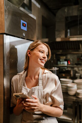 Young blonde woman using cellphone while working in restaurant kitchen