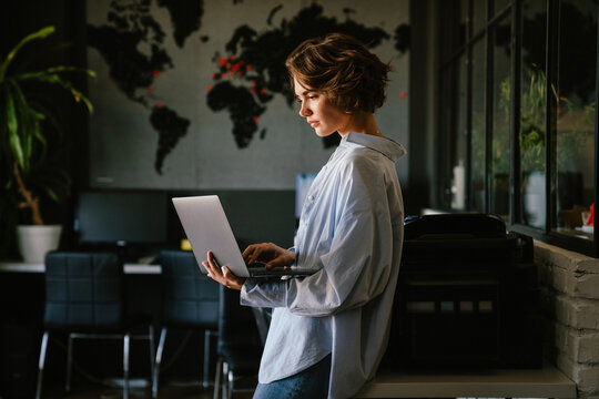 Concentrated business woman using laptop while standing in modern workspace