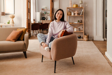 Beautiful asian woman smiling at camera while sitting on armchair at home