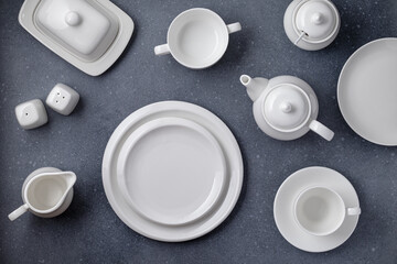 Obraz na płótnie Canvas White tableware for serving and eating meals. Empty clean plates, cup, teapot, sugar bowl, milk jug and butter dish on a gray concrete table, top view. Flat design. Ceramic crockery. Chinese tea set.