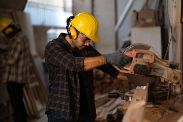 Young carpenter wears safety goggles, earmuffs and yellow helmet, use a circular saw to cut small...