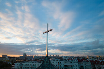 A cross on top of the roof of a Catholic Church in Warsaw, Poland is seen against the backdrop of a blue sky with a few clouds during sunset. 