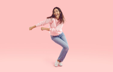 Laughing brunette woman in jeans and pink sweatshirt dancing cheerfully and singing isolated on pink background, full length portrait. Disco, dance, sing, banner for advertisement and marketing.