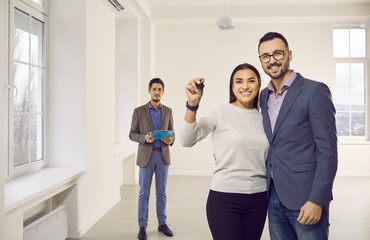 Young family couple buying or renting new house or apartment. Portrait of happy man and woman showing key while standing together in white living room at home, with real estate agent in background
