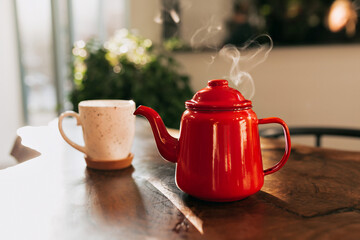 Stylish red kettle with steam with cute white cup slanting on wooden table in cafe in sunlight. Cup of hot tea. Tea or coffee on the table in sunny day 