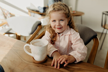 Wonderful happy excited girl with curly hair wearing pink shirt sitting in modern sunny cafe with big cup of cacao and smiling at camera 