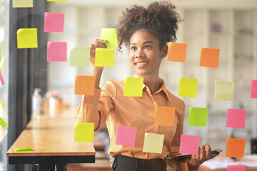 Positive African American female employee putting sticker notes to share idea for startup project on glass board in office