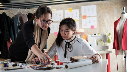 Asian middle-aged female fashion designer teaches a young teen trainee tailor in studio with colorful thread and sewing fabric for dress design collection ideas, professional boutique small business.