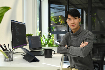 Young male website developer sitting front of computers with coded data on screen and smiling to camera