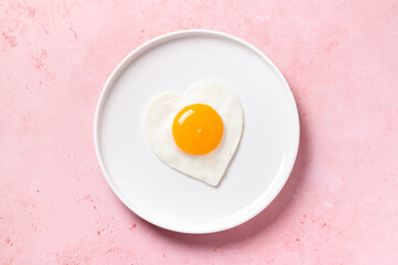 Fried egg in heart shape on white plate, romantic food concept, pink background