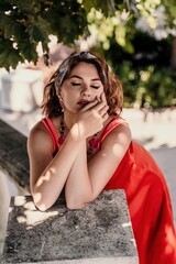 A pretty woman in a red silk dress and a bandage on her head smiles against the background of the leaves of a tree. She is leaning on the coop and looking into the camera. Vertical photo.