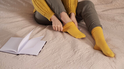 unrecognizable woman puts on cotton warm yellow sock on bed closeup. create comfort at home concept