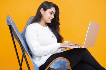 Young indian girl wearing white T-shirt using laptop while sitting on chair isolated over orange...
