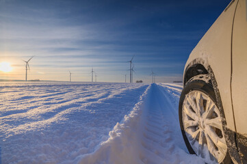 Car tires on winter road covered with snow. Vehicle on snowy country road with wind turbine in the...