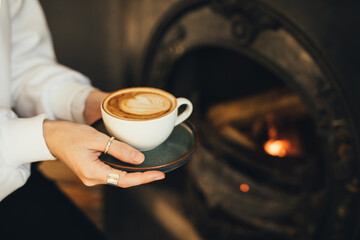 Close up female hands holding cup of hot cappuccino on wooden stool near burning fireplace.