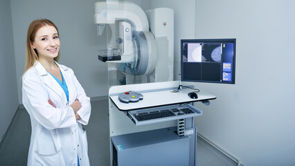 Mammography, mammogram. Positive medical technician with arms crossed standing near mammography workstation with imaging machine for breast screening - obrazy, fototapety, plakaty