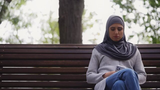 Unhappy muslim woman in hijab sitting alone on bench in park, feeling sadness