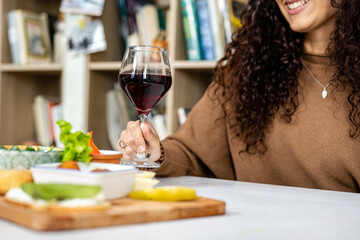Close up of a glass of red wine held in hand by a smiling young woman while eating an assortment of vegetables and vegan foods