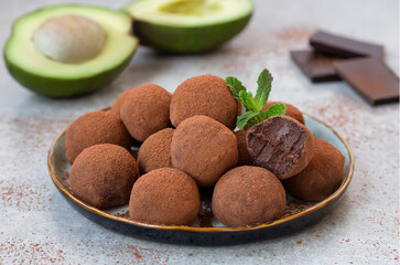 Dark Chocolate Avocado Truffles.  Assorted chocolate candy balls rolled in cocoa powder served on a plate. Sliced avocado and chocolate pieces on background. Selective focus, horizontal.