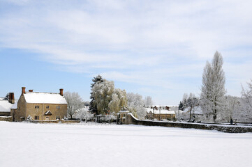 Manor house in the Cotswolds covered in snow with a village behind,  Blue sky and clouds.  poplar trees and a willow tree.