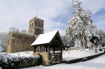 A church in Oxfordshire  from below in the sunshine and snow with the lychgate in foreground.  Blue sky and clouds behind