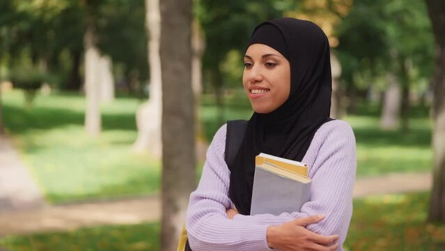 Cheerful arab female student holding books and smiling while resting campus park