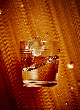 Glass Tumbler Falling With an Ice Cube and Water on a Wood Grain Background